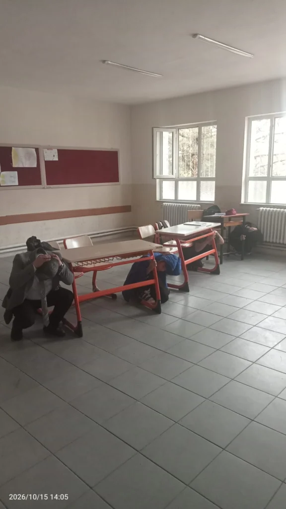 Tatbikat sırasında öğrenciler sınıfta deprem anı uygulaması yaptı.
Öğrenciler sınıfta sıraların yanında başlarını koruyarak tatbikata katılıyor.